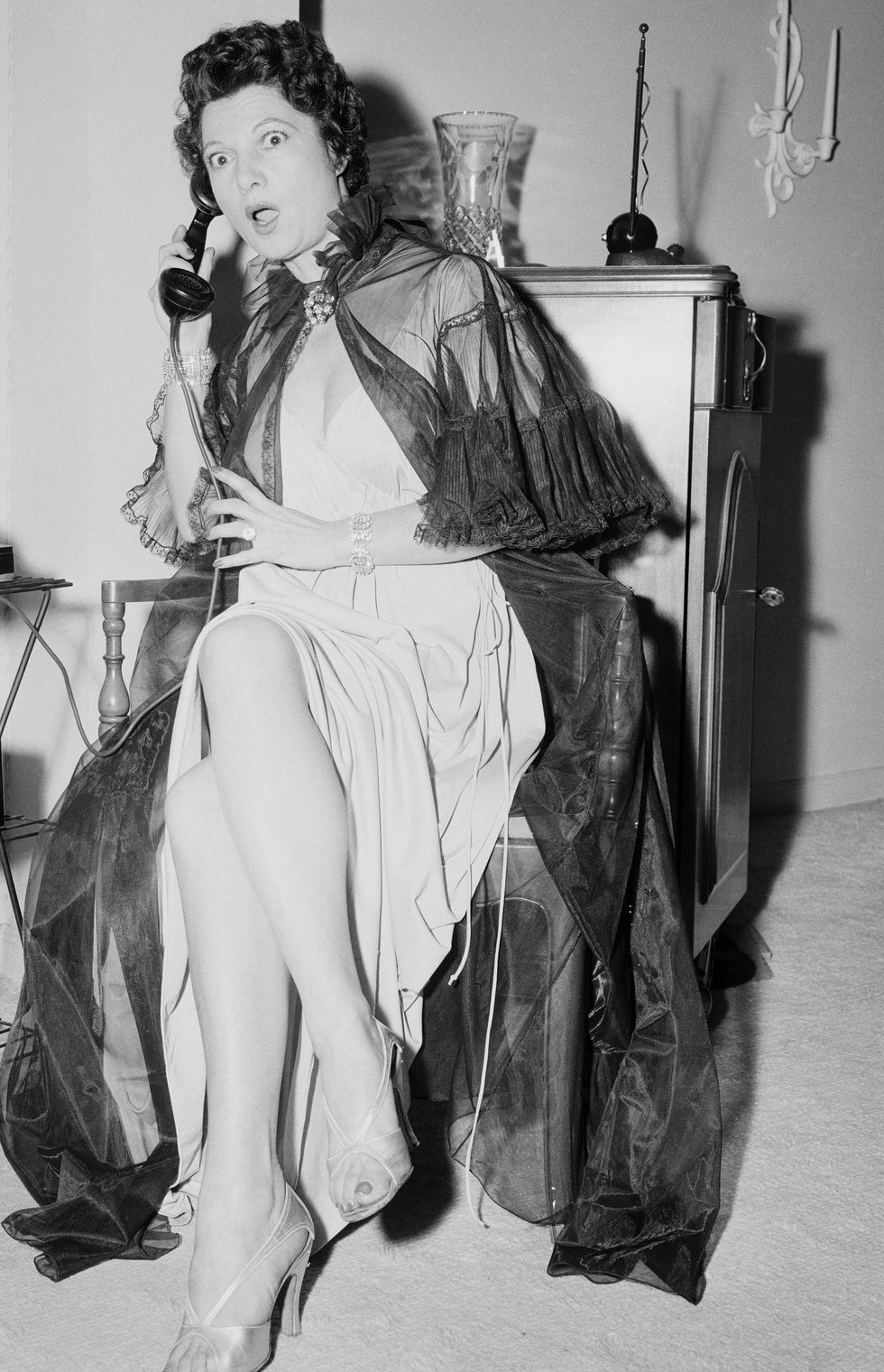 A woman in theatrical garb appears shocked by what she is hearing over a telephone.