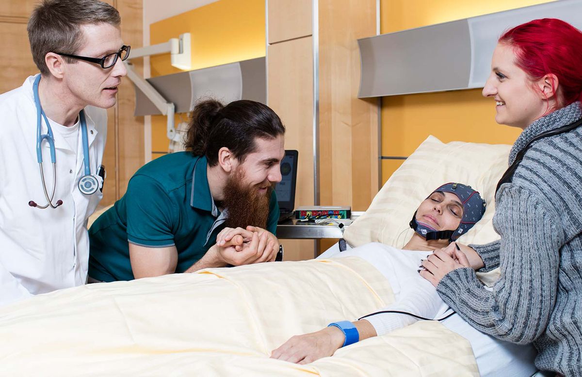 A woman lies in a hospital bed with the mindBEAGLE EEG headpiece on, surrounded by two family members and a doctor.
