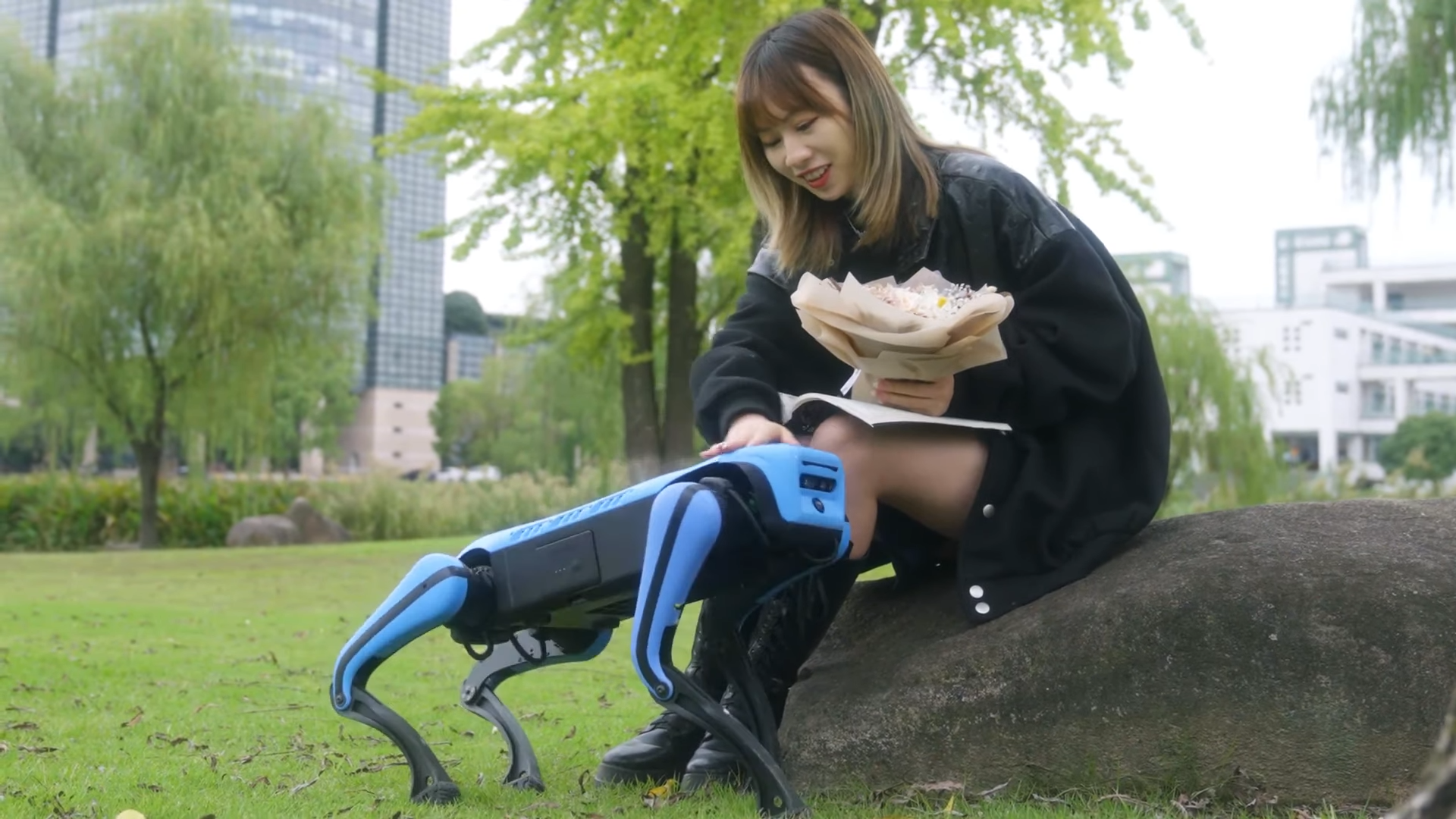A woman sitting on a rock and holding flowers pets a blue and black robotic dog