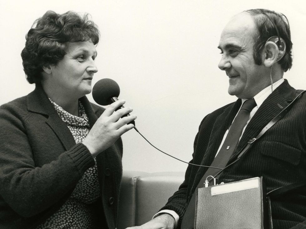 A woman speaks into a microphone connected to a book-sized box that is in turn connected to his ear.
