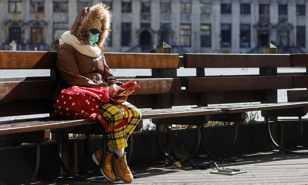 A woman wearing a facemask for protective measures looks at her mobile phone as she sits alone on a bench at the Grand-Place in Brussels, on March 18, 2020 as a strict lockdown came into in effect in Belgium to stop the spread of COVID-19, caused by the novel coronavirus.