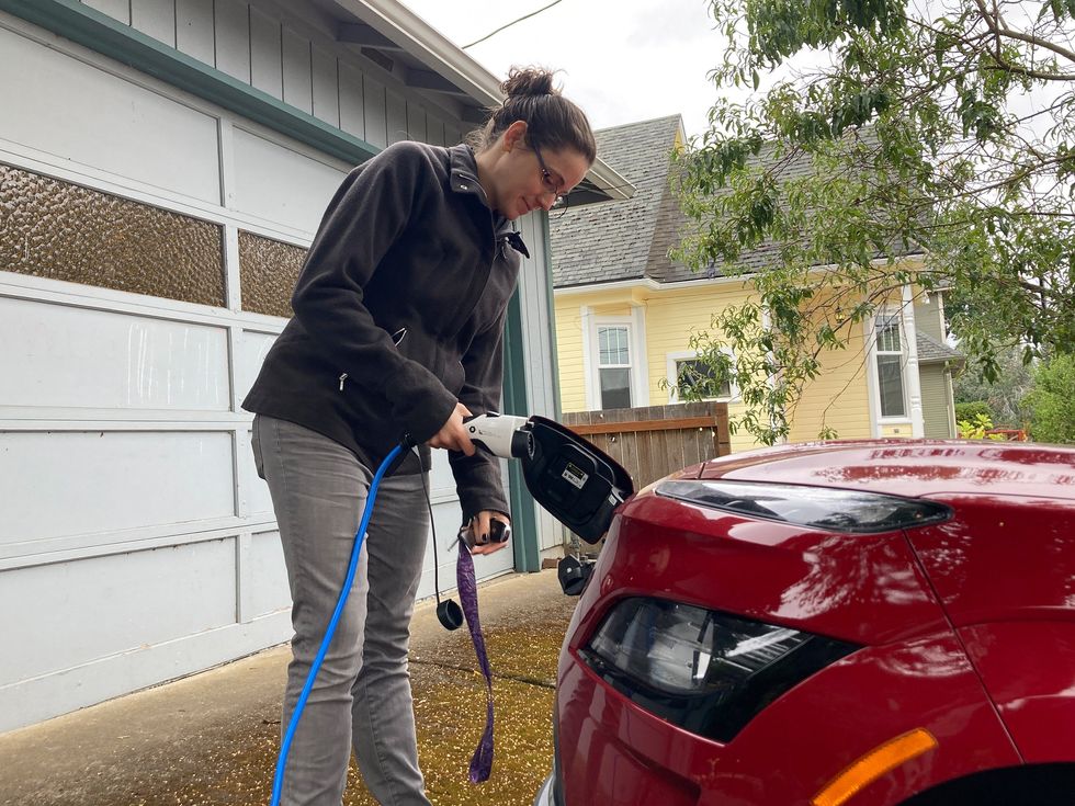 A woman wearing grey jeans and a jacket charges a red electric vehicle in front of a residential garage.