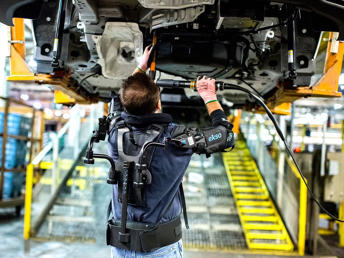 A worker on a car assembly line works on the chassis above his head. He's wearing an upper-body exoskeleton that has components around his arms and down his back.