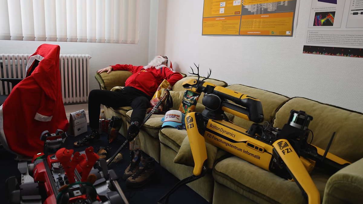 A yellow legged robot wearing antlers is passed out on a couch in an office next to Santa Claus.