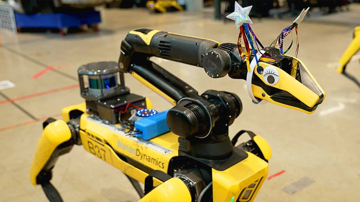 A yellow robot dog has a black and yellow arm mounted on its head, where the gripper of the arm is decorated with eyes and colorful ears.