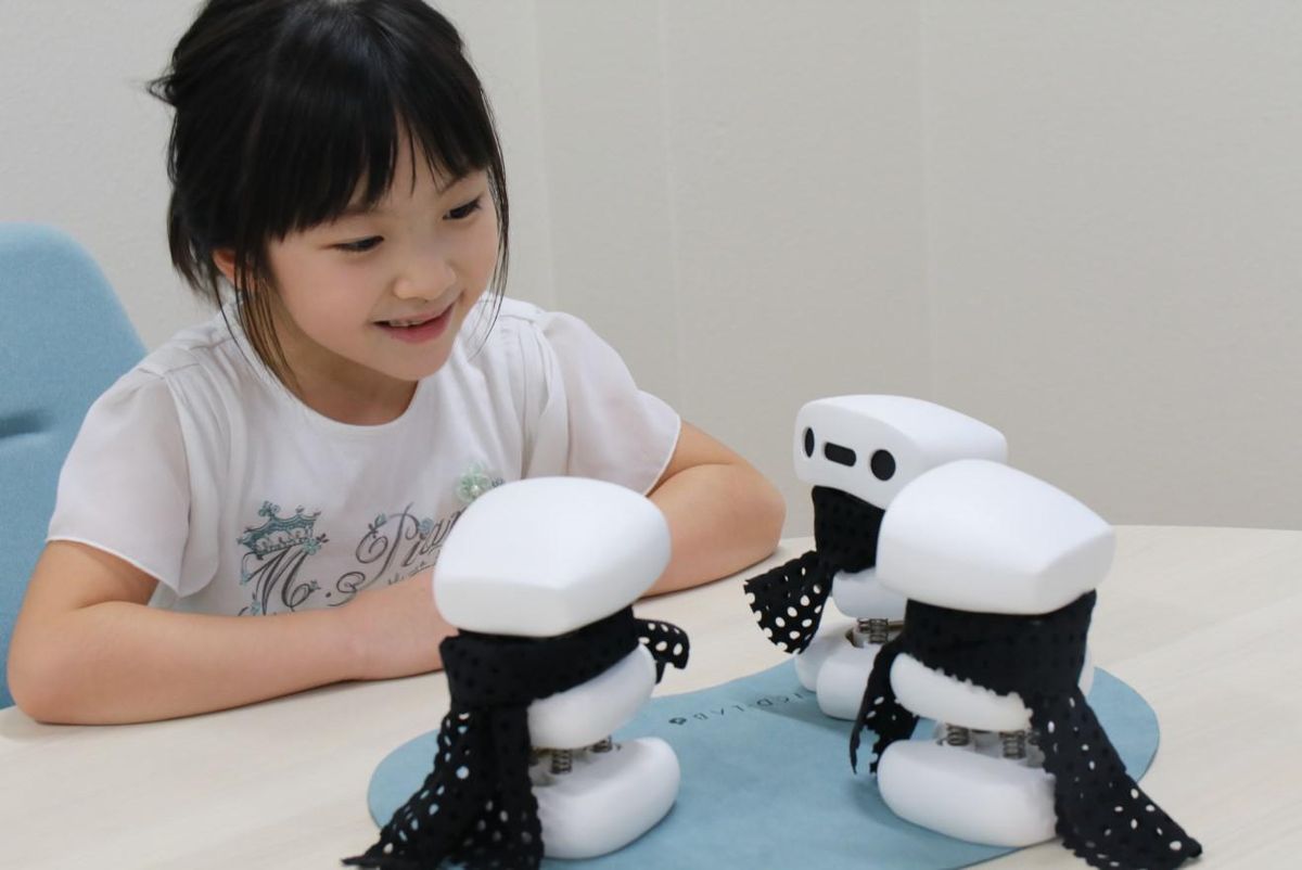 A young girl looks at a cluster of three simple robots facing each other on a table