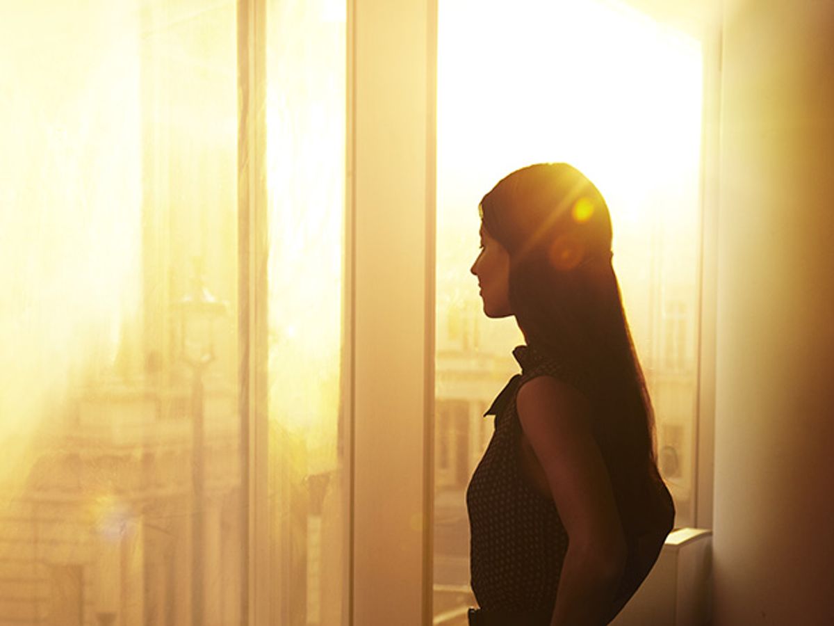 A young woman looks out of a large office window towards a group of neoclassical buildings. The scene is tinted yellow by light from a sun low above the horizon.
