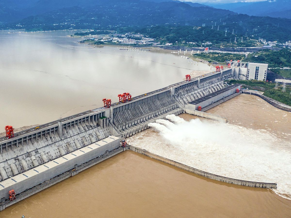 An aerial view of a long, concrete dam, the farther end of Yangtze River, which rests on the bank of a river, at which point there is a white building. In the middle of the dam, at its base, three white jets of water are spouting white, turbulent water into a holding area. 