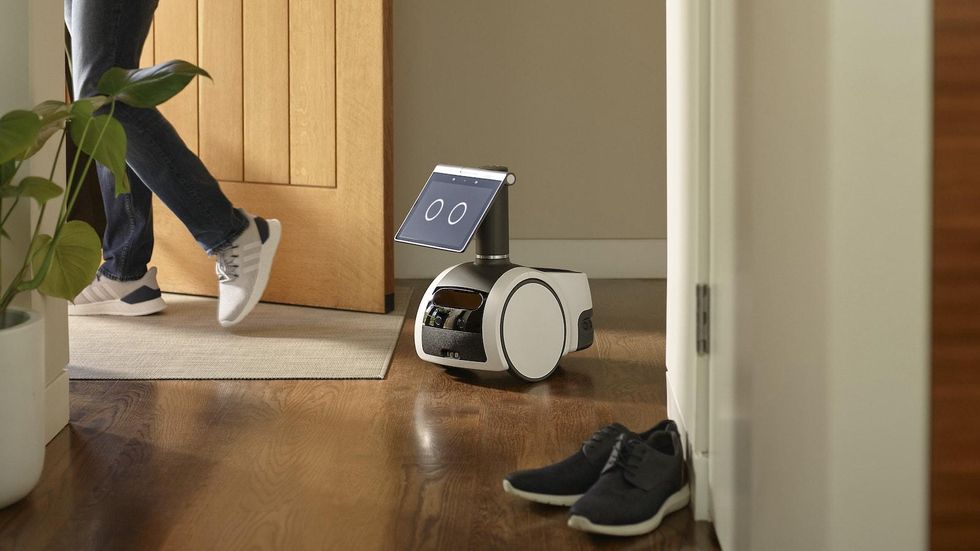 An Amazon Astro robot, with two wheels and knee-high body with a tablet screen with two circles as eyes, stand next to a person who is answering the door.