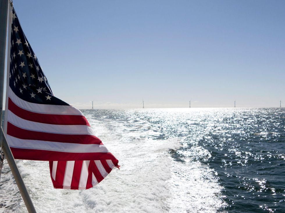 An American Flag in the foreground with water and wind turbines in the distance.