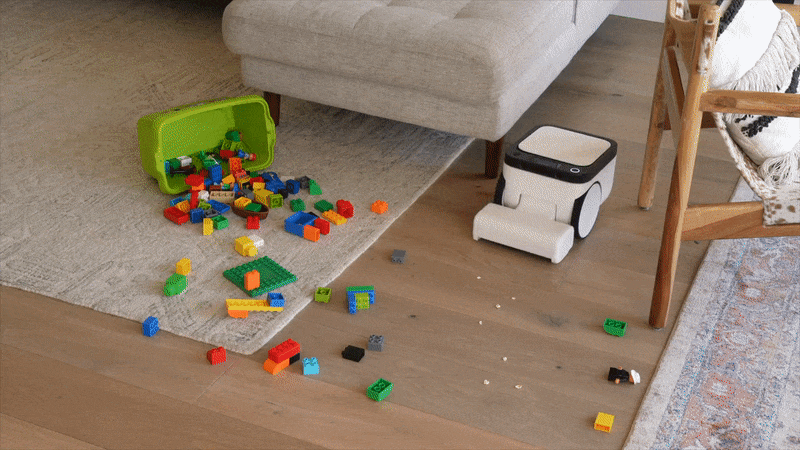 An animated GIF showing a boxy black and white robot vacuum cleaning a floor while navigating around spilled LEGO bricks