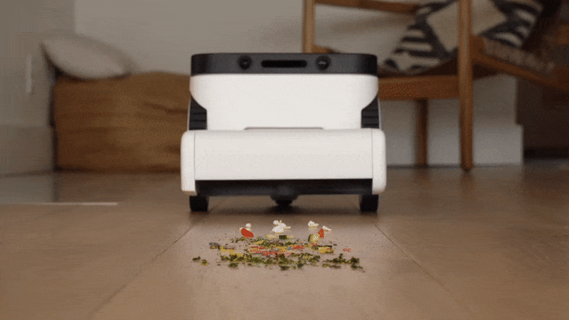 صورة متحركة بتنسيق GIF تُظهر عرضًا لمستوى الأرض لمكنسة روبوت أبيض وأسود يشبه الصندوق ينظف الفوضى