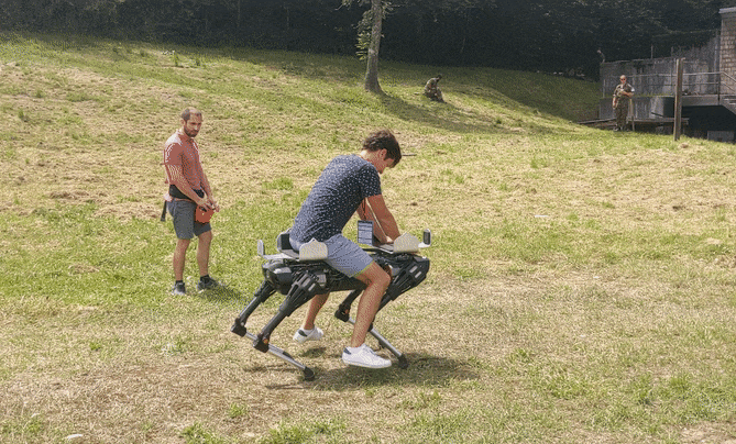 An animated GIF showing a human riding on top of a small quadrupedal robot on a grassy field.