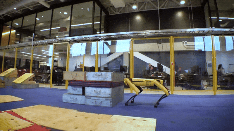 An animated GIF showing a yellow and black robotic dog jumping onto a stack of boxes in a research lab.