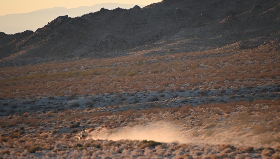 An ATV kicks up dust as it drives off into the distance of a desert landscape near sunset