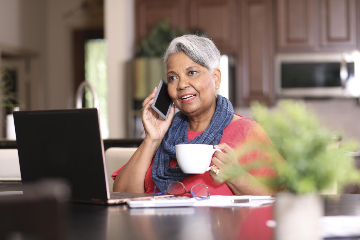 An elderly woman at home talks on the phone in front of a computer, holding a cup of coffee