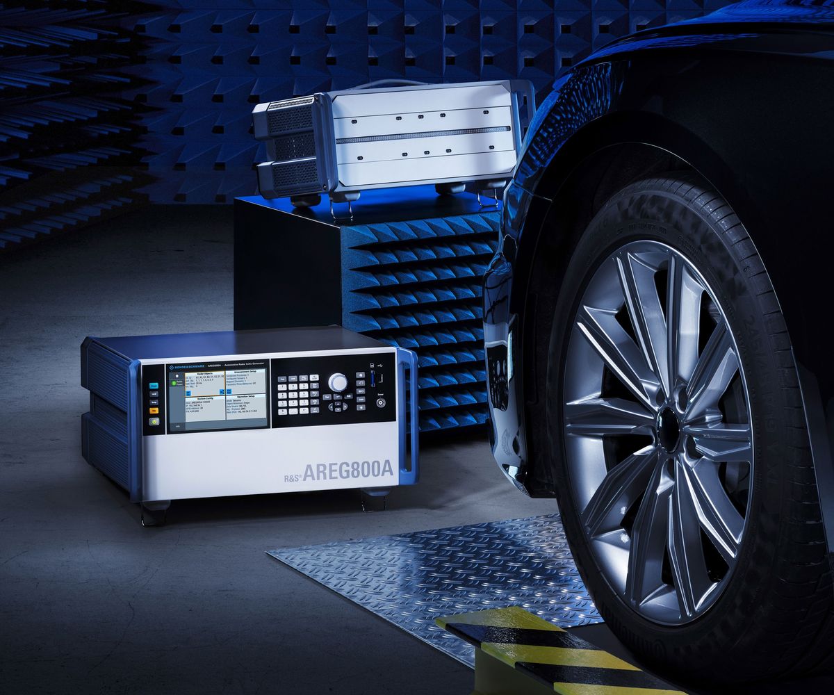 An electronic radar echo generator from Rohde & Schwarz the size of shoebox with buttons and an LCD screen sit on the floor next to the wheel of a car with background walls covered in sound absorbing foam tiles.
