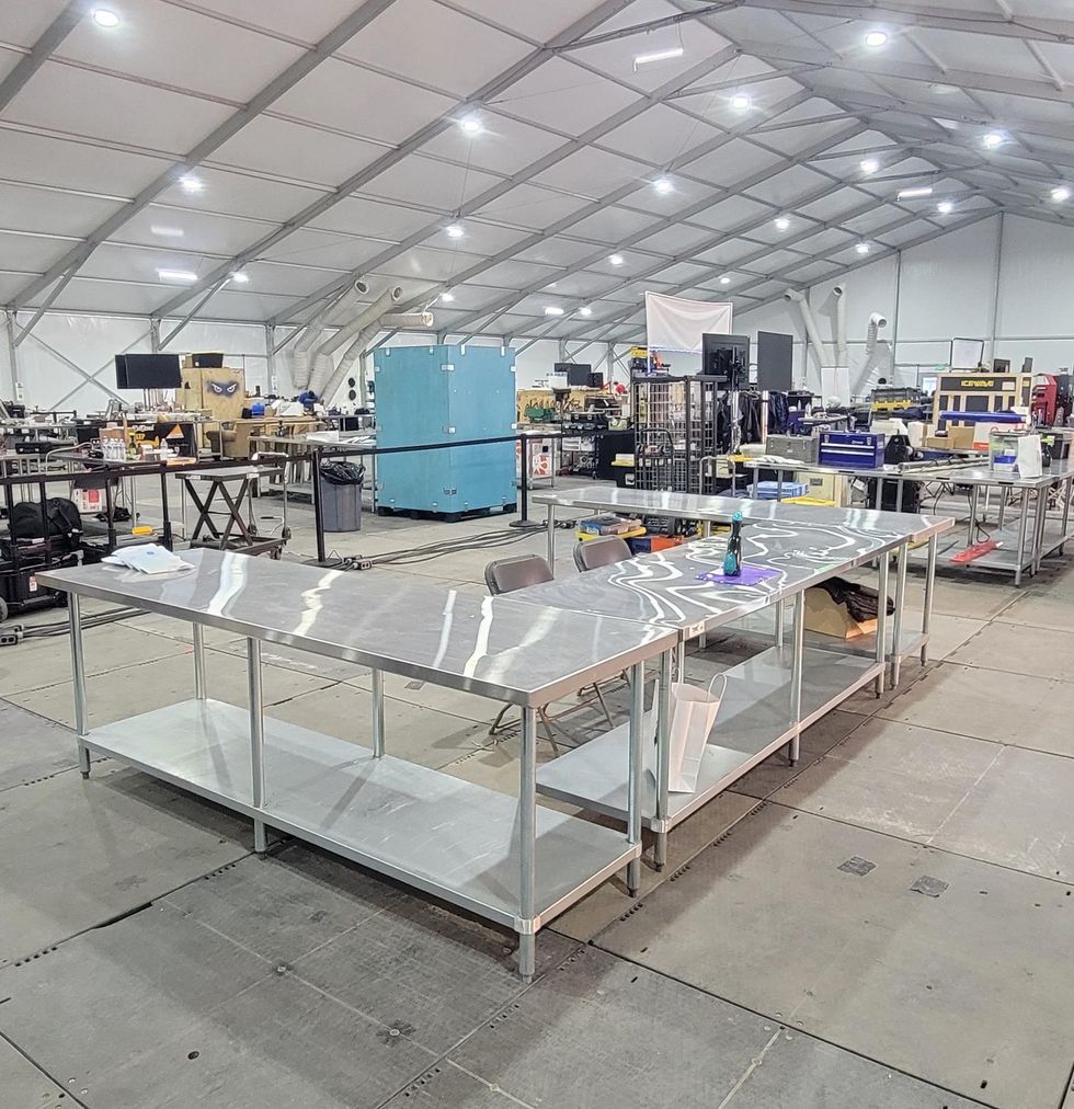 An empty U-shaped set of metal workbenches in a large tent crowded with workspaces