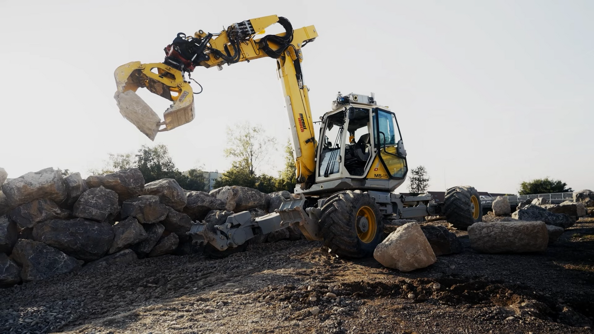 An excavator with no human inside and sensors on its roof picks up a large rock and places it on a partially completed rock wall.