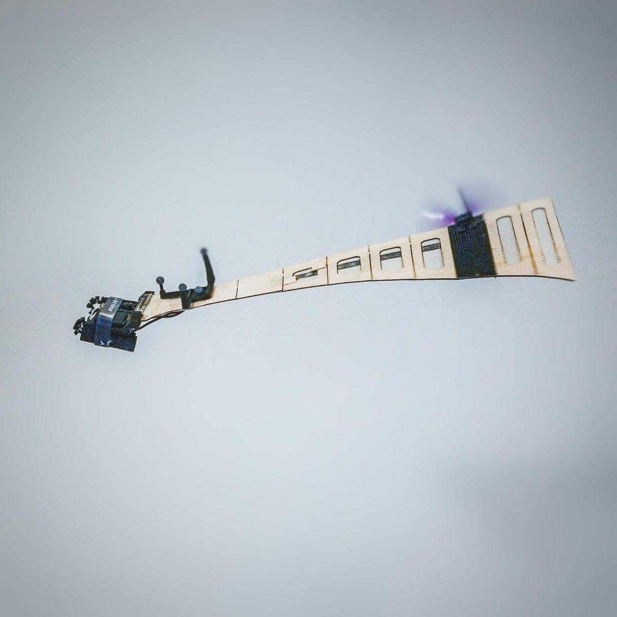 An in flight picture of a single wing drone made out of balsa wood with batteries on one end and a propeller on the other