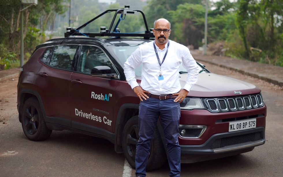 An Indian man in glasses stands with his hands on his hips in front of a car labeled driverless car and RoshAi.
