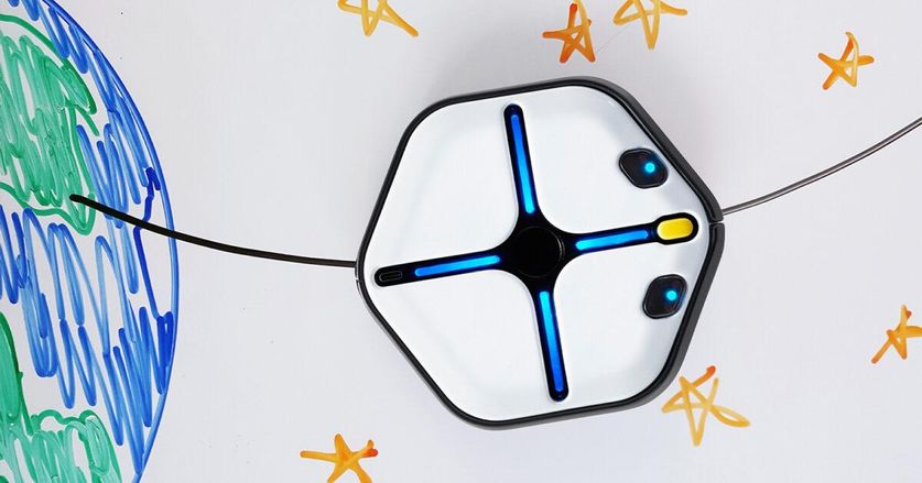 An iRobot Root, with a hexagonal white body with blue and yellow lights, uses its magnetic wheels to drive vertically on a whiteboard that has planet Earth and stars drawn on it.