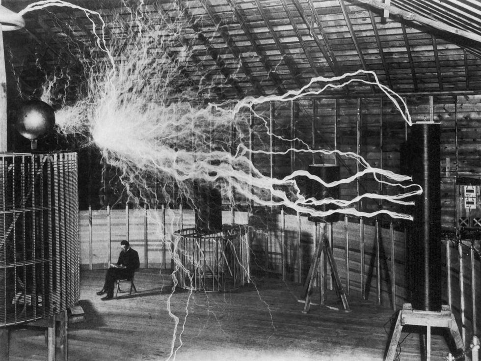 An old black and white photo showing a man sitting next to a large electrical apparatus that is emitting sparks.