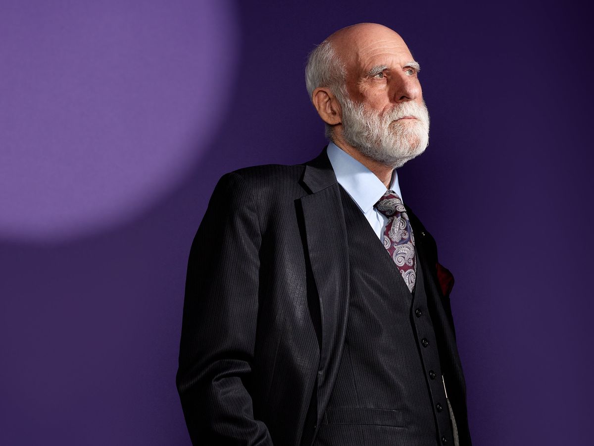 An older man with white hair wears a suit, and looks off into the distance as he leans against a purple background.