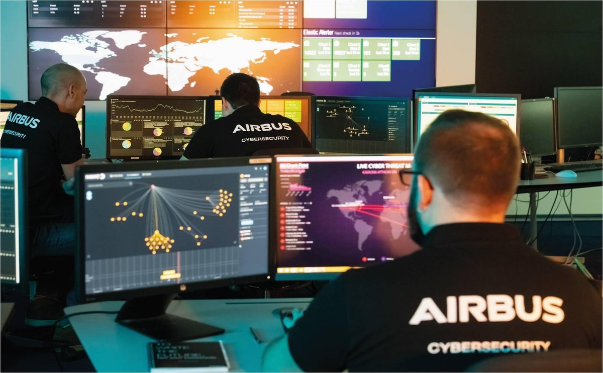 An operations center with seven computer screens and a big screen on the wall, with three people monitoring those screens, with the back of their shirts reading “Airbus Cybersecurity"