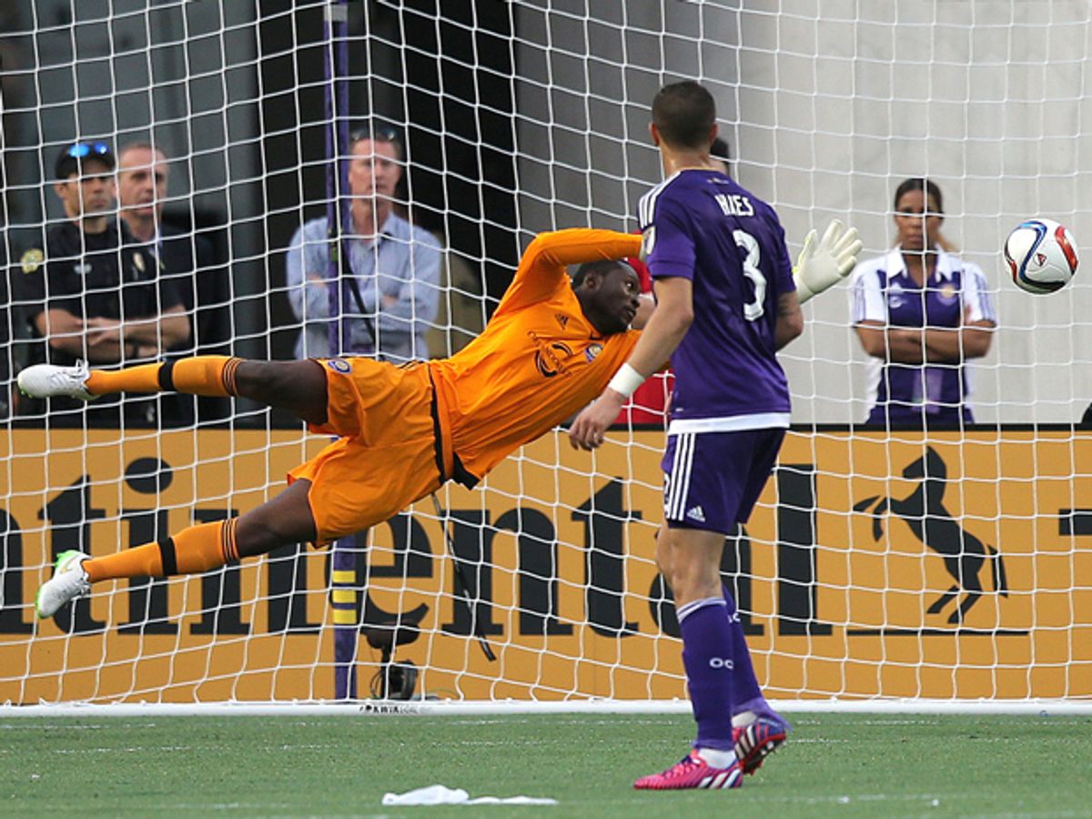 An orange-clad goalkeeper dives towards a soccer ball, while another player in a purple uniform watches