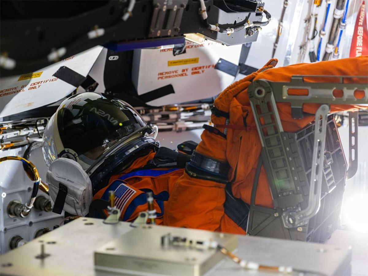 An orange survival space suit reclines inside of a a cramped space capsule