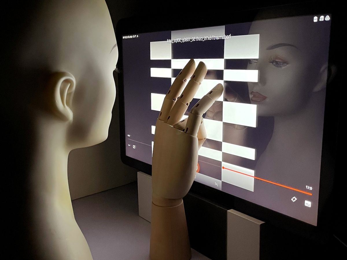 An photograph of a mannequin head and hand placed in front of a computer screen that is displaying a checkerboard-like pattern