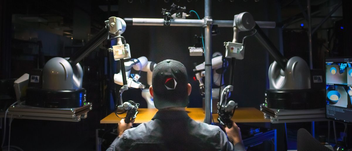 back view of man in baseball cap holding controllers for robot arms, which are in silhouette in front of him, just outside of the shadows