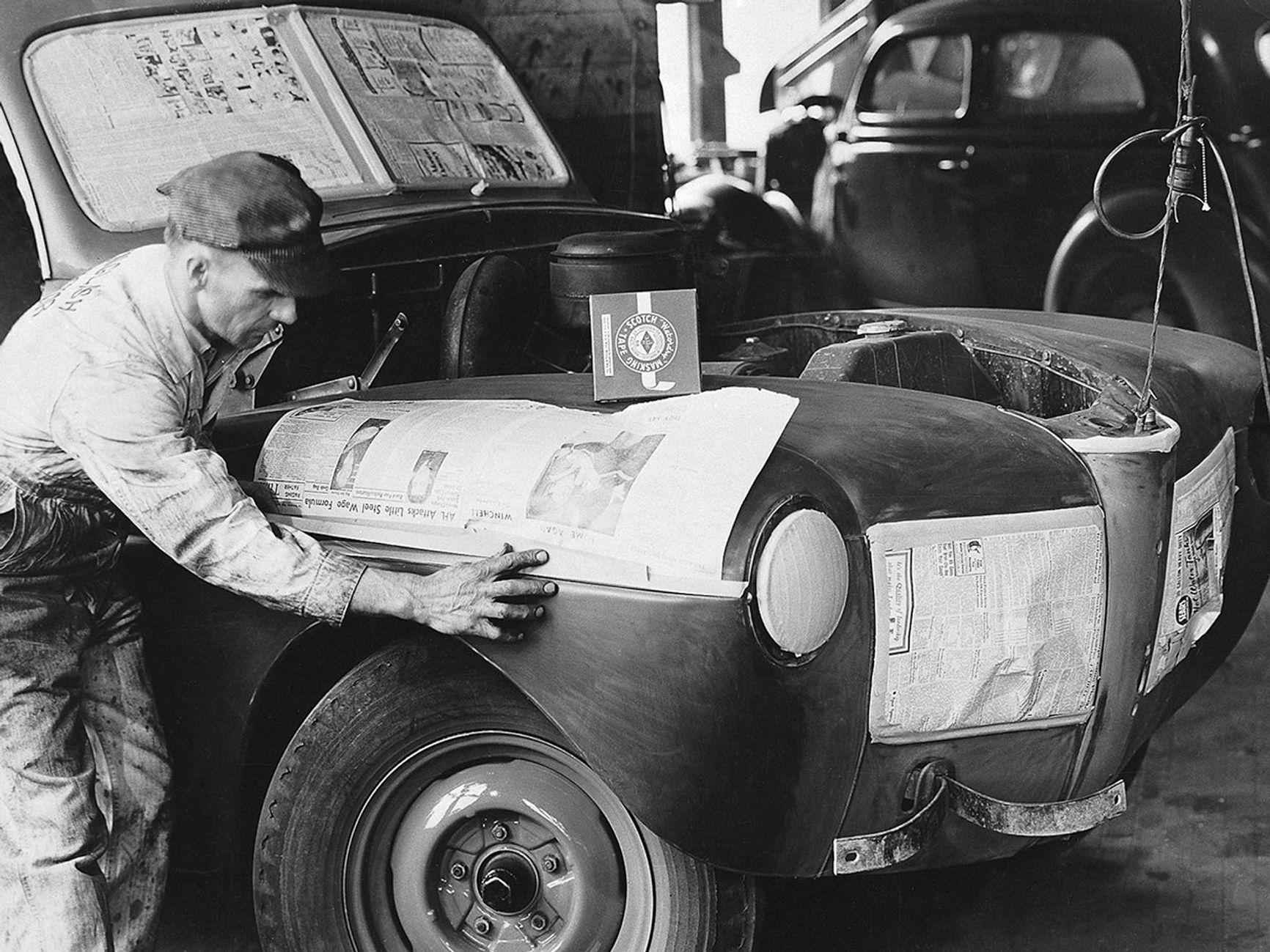 Black and white historical photo of a man in worker overall and a cap applying newspaper and tape to an old car.