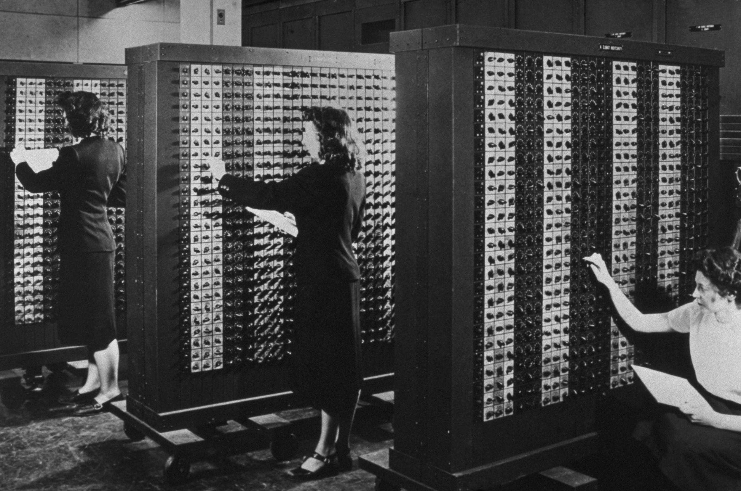 Black and white image of three women, each in front of a large wheeled cabinet faced with countless rotary switches