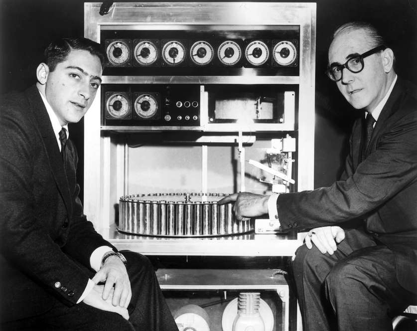 black and white image of two men sitting on either side of a large mechanical box with knobs and a cylinder plate on the bottom