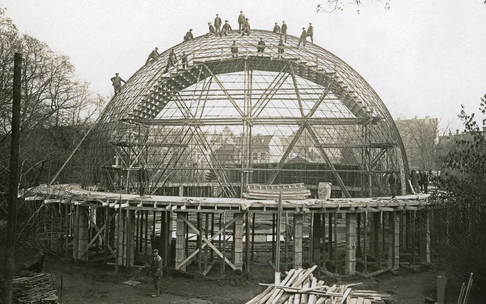 Black and white photo of a domed structure under construction with workers standing on the top.