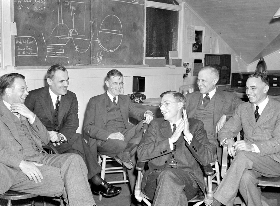 Black and white photo of a group of six seated men smiling and laughing in front of a chalk board with scientific drawings