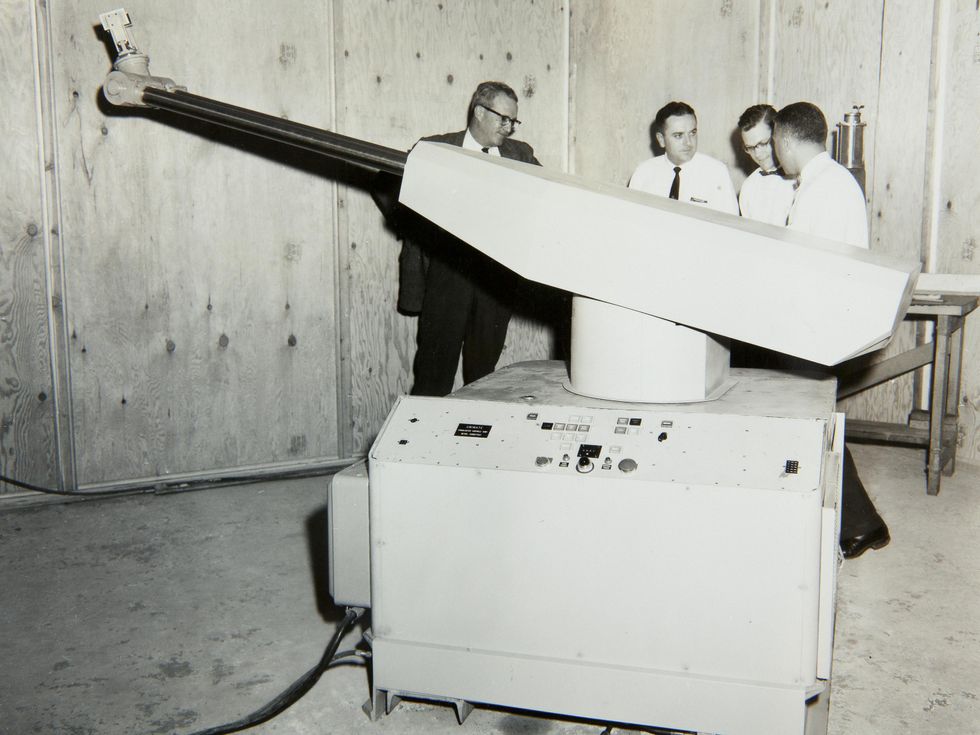 Black and white photo of a large robotic arm on a massive base that is almost as tall as the 4 men standing behind it.\u00a0