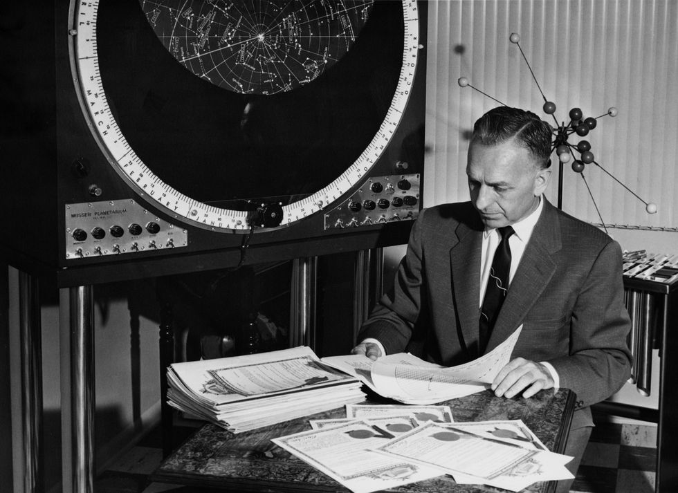 Black and white photo of a man in a suit, sitting at a desk in front of a round display. 