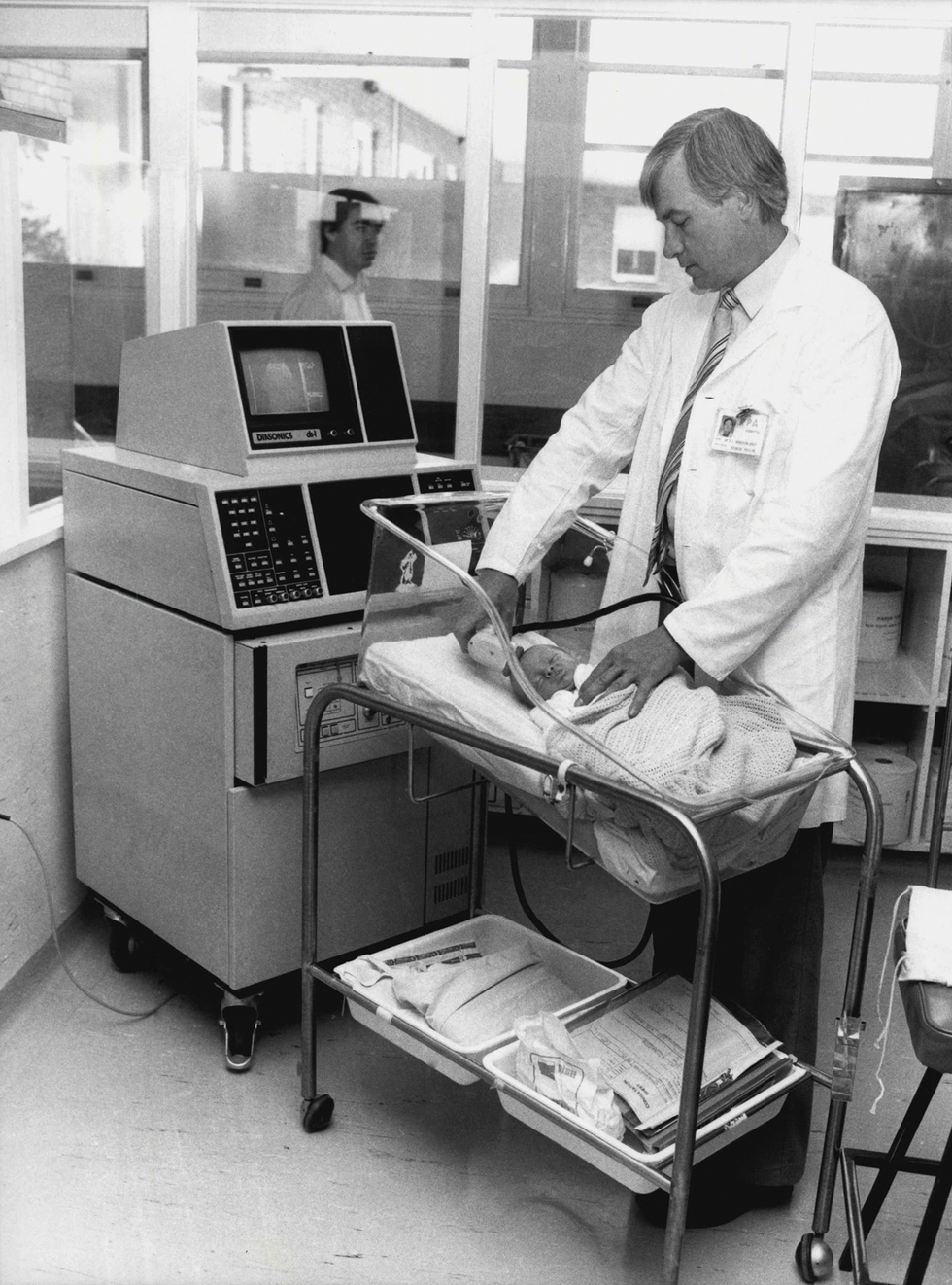 Black and white photo of a man taking an ultrasound image of a baby in a crib using old, bulky ultrasound equipment