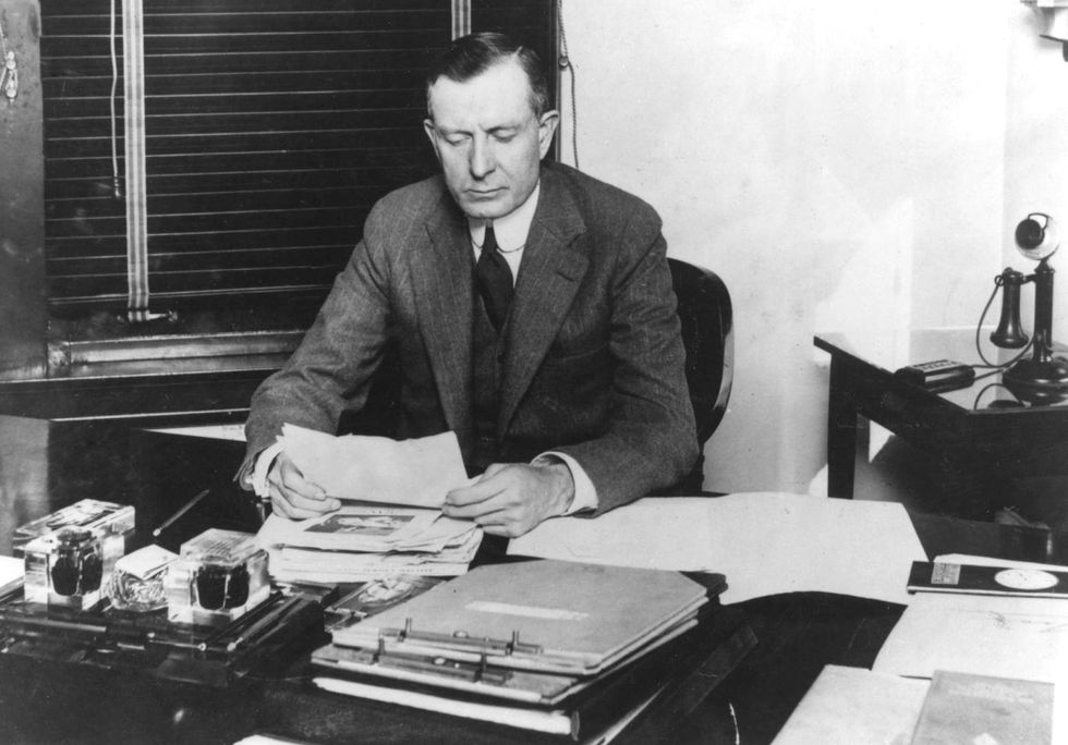Black and white photo of a middle-aged white man in a suit sitting at his desk and looking at papers.