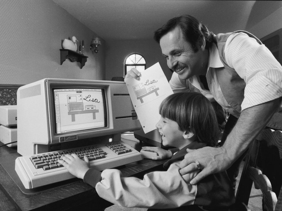 Black and white photo of a smiling man looking on as a young boy types at a computer keyboard.