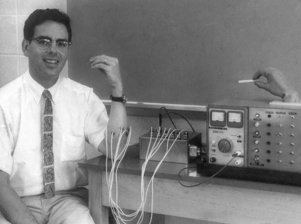 Black and white photo of a smiling young man whose left forearm is attached by wires to electrical equipment. 