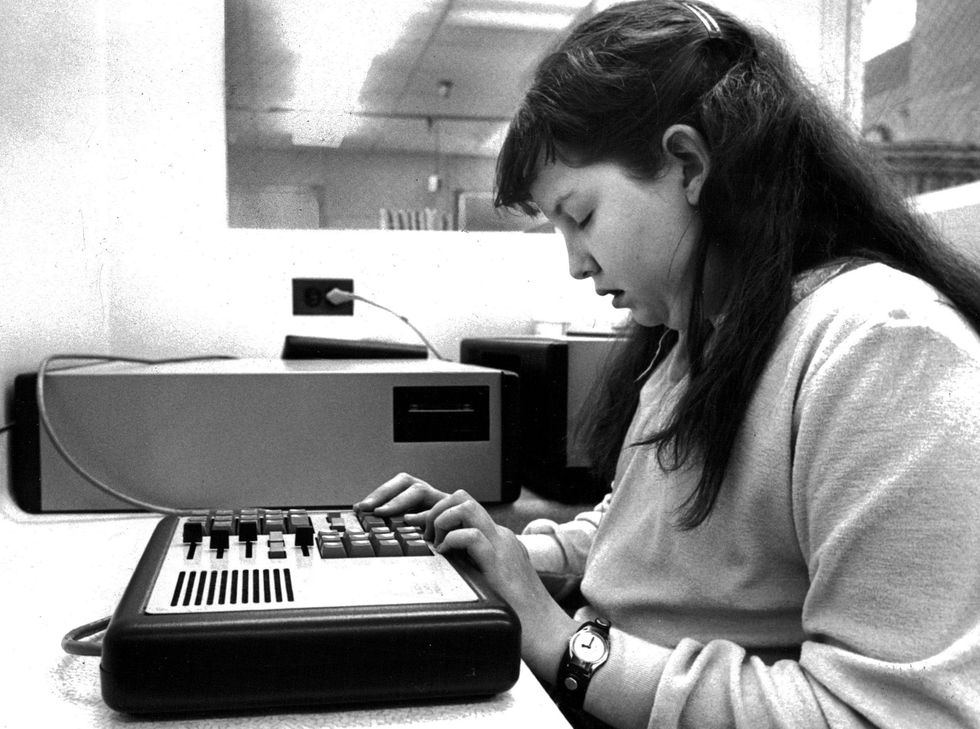 Black and white photo of a young dark haired girl with her eyes closed, and her fingers resting on a rectangular machine with buttons on it.