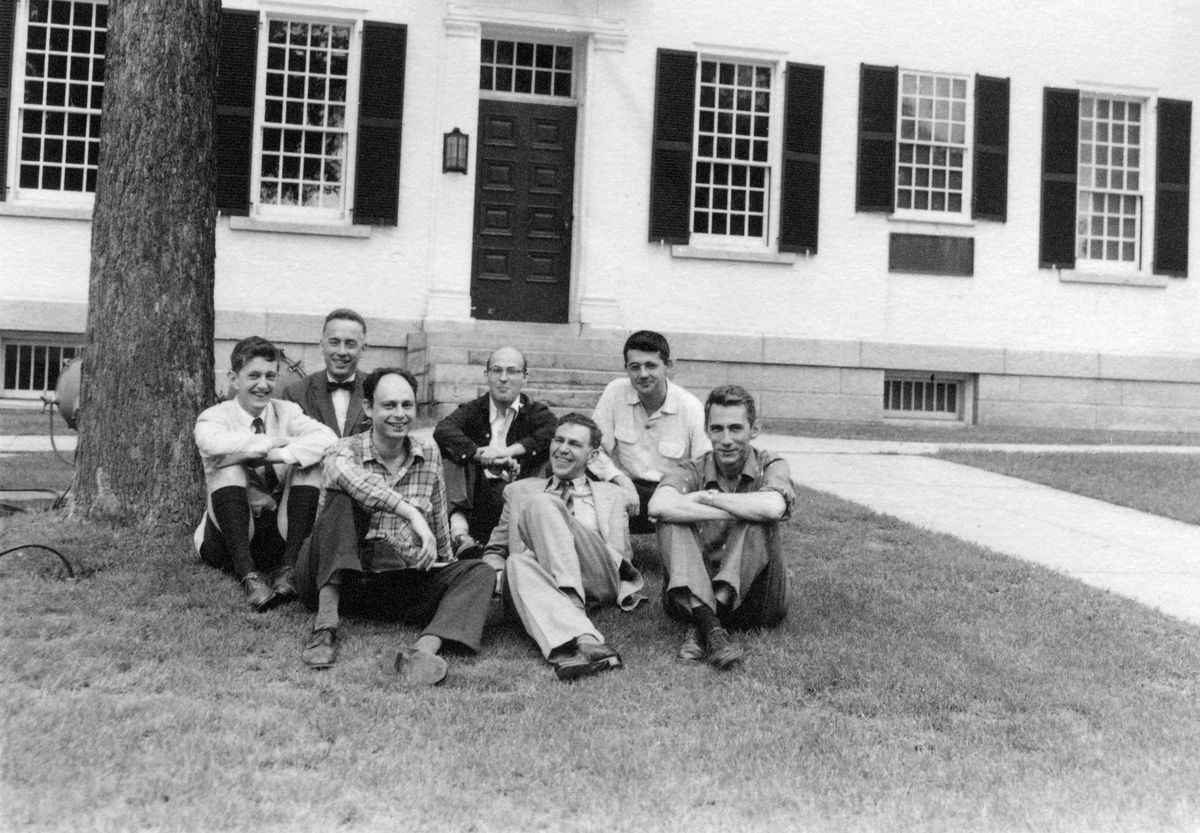 black and white photo of seven smiling men, sitting on a lawn in front of a tree and a white school building with many windows.