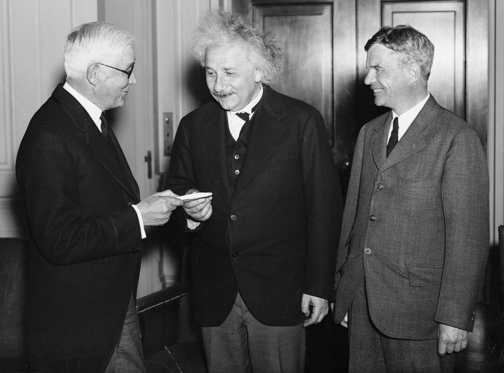 Black and white photo of three smiling men in suits. The man on the left is handing something to the man in the middle, while the man on the right looks on.