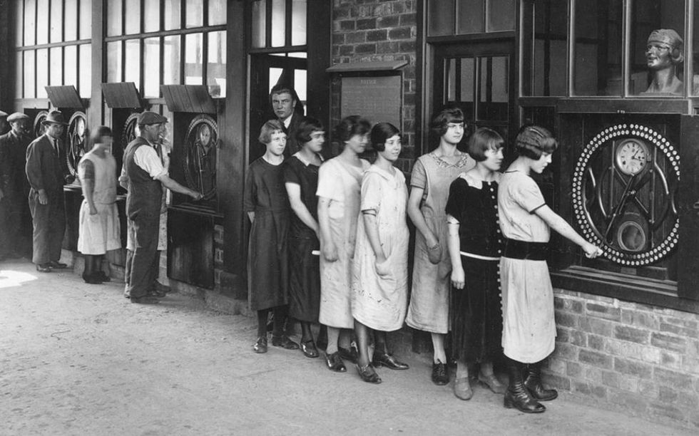 Black and white photo of women standing in line and watching as one woman turns the handle on a large dial. 