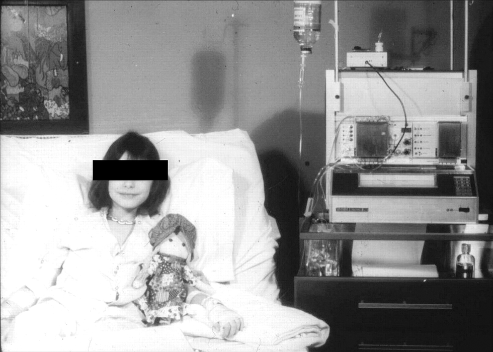 Black and white photo shows a young woman, her face blacked out, with a doll in bed. She is hooked up to a large machine on the right.