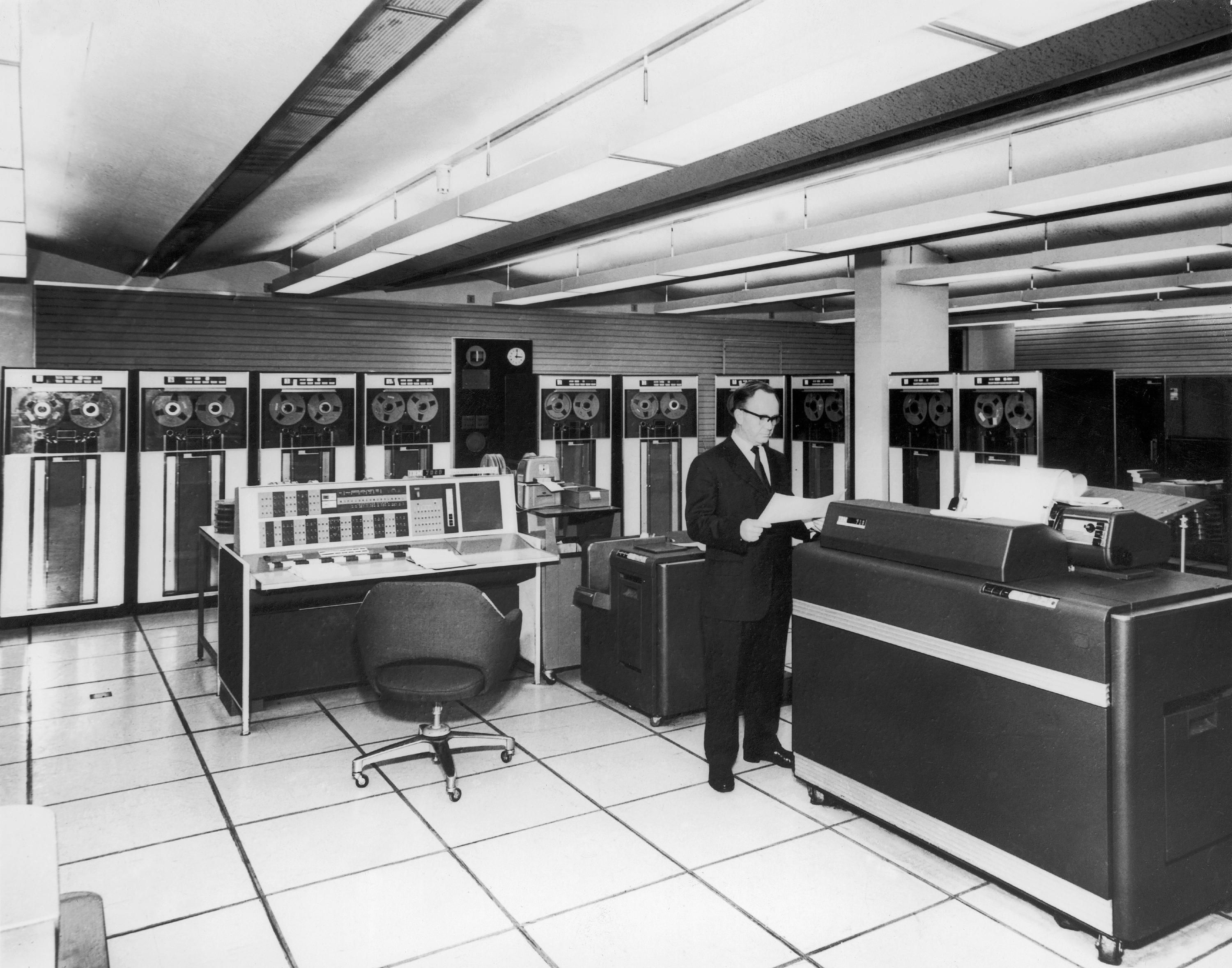 Black and white photograph of a man in a large room with mainframe computer elements lining the walls